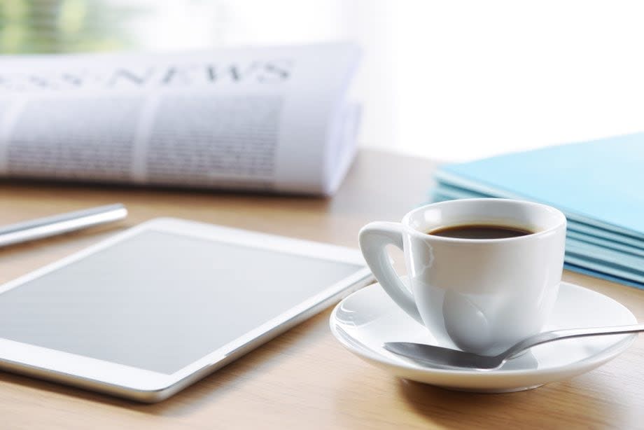 Ein Zeitung, ein Tablet, ein Stapel Blätter und ein Kaffeetasse mit Untersetzter und Löffel stehen auf einem Holztisch.