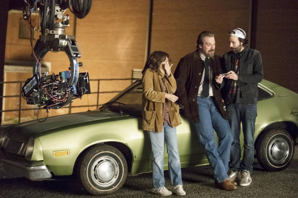 The three of them leaning against a car as Winona covers her face and David laughs while looking at the phone