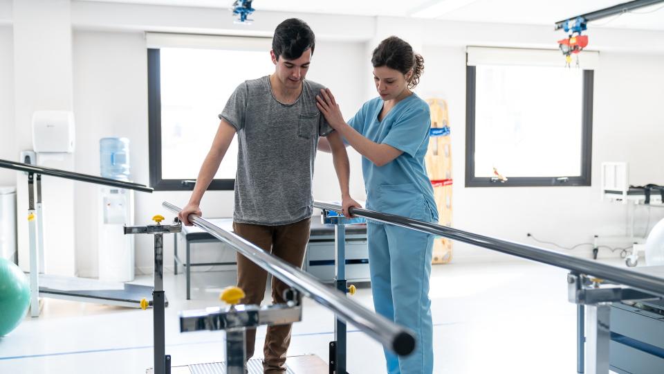 Focused male patient at physical therapy walking with the help of parallel bars and therapist next to him giving support.