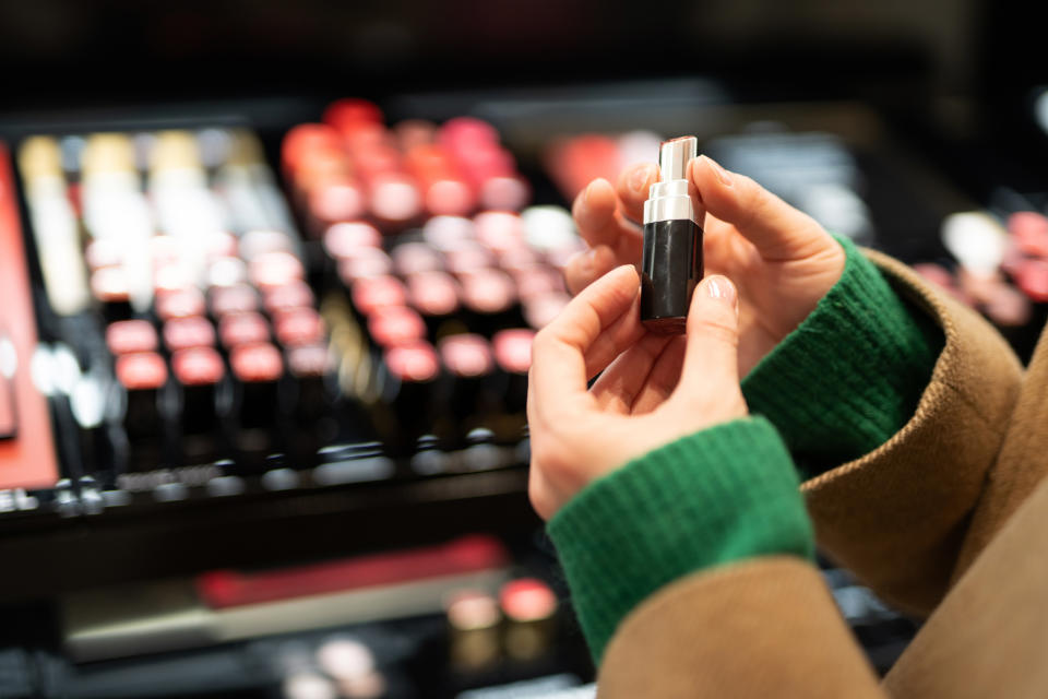 Person holding a lipstick in a store with many lipsticks in the background, examining it closely