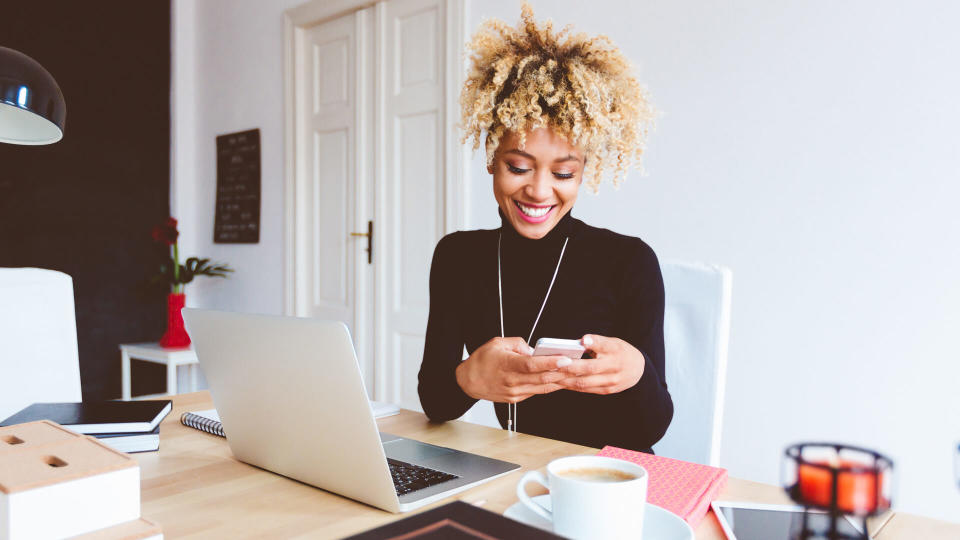 Beautiful afro american young woman sitting at the desk in a home office and using a smart phone.