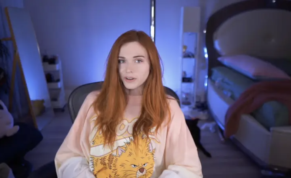 A woman with long red hair in a pink sweatshirt sits in a darkly lit room looking at the camera