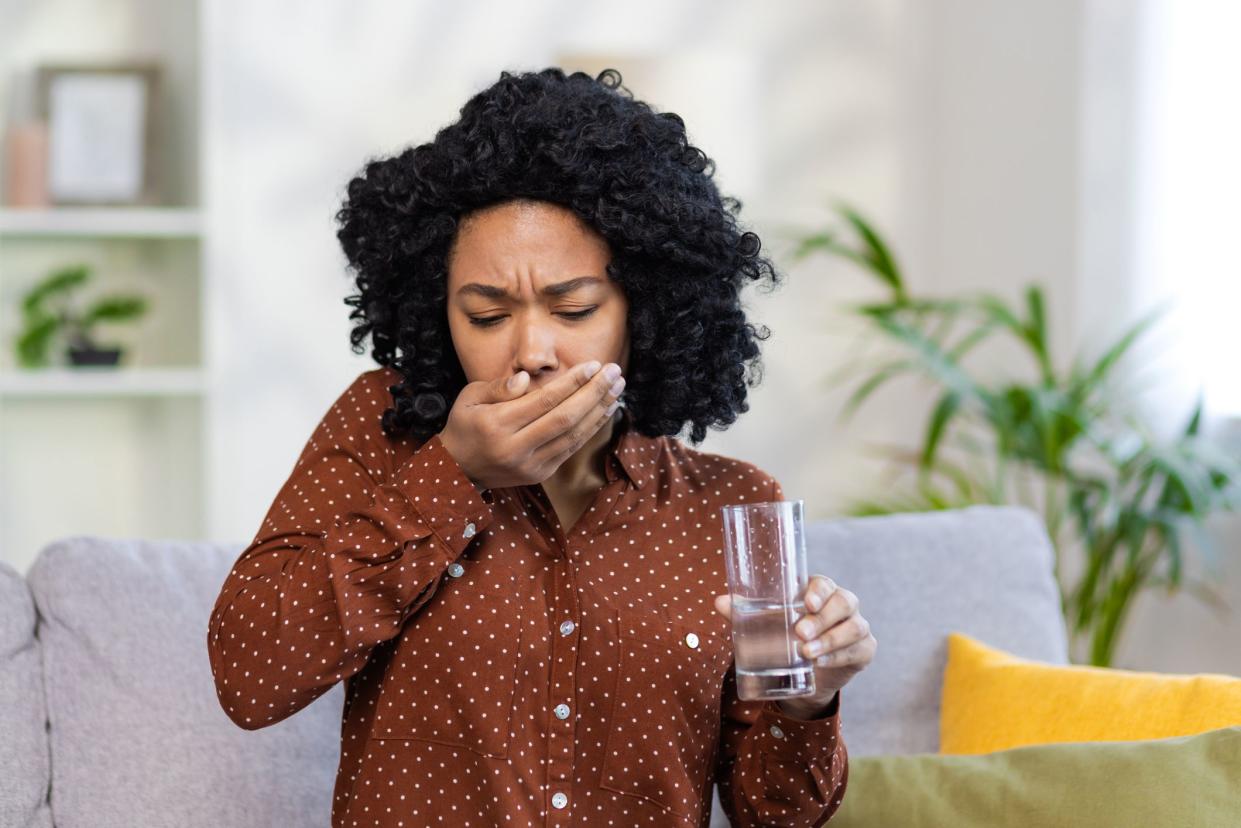 A sick young African-American woman sits at home on the sofa and drinks pills, holds a glass of water in her hands, feels bad.