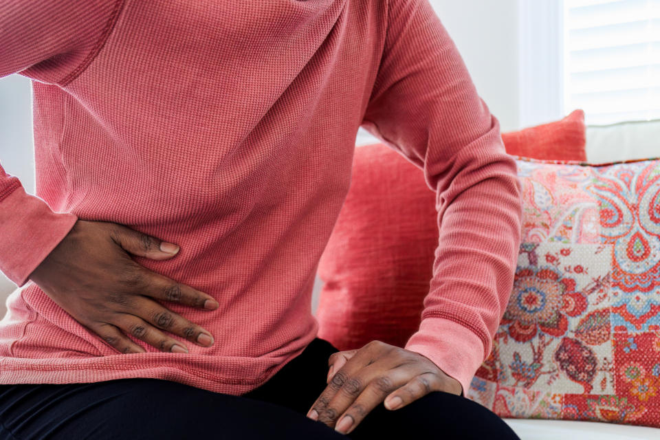 A person wearing a long-sleeve shirt holds their side, appearing to experience pain, while sitting on a couch with patterned pillows