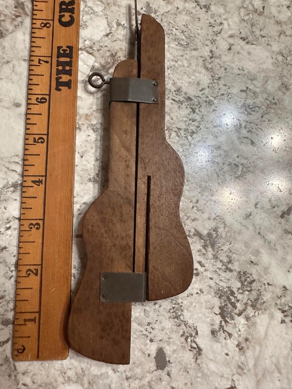 A wooden puzzle piece in the shape of a guitar next to a scale ruler. The puzzle has a metal clasp and hinge