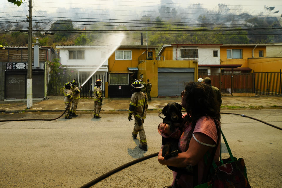 A woman evacuates with her dog as firefighters conduct a protective water spray on homes as forest fires burn nearby, in Vina del Mar, Chile, Saturday, Feb. 3, 2024. Officials say intense forest fires burning around a densely populated area of central Chile have left several people dead and destroyed hundreds of homes. (AP Photo/Esteban Felix)