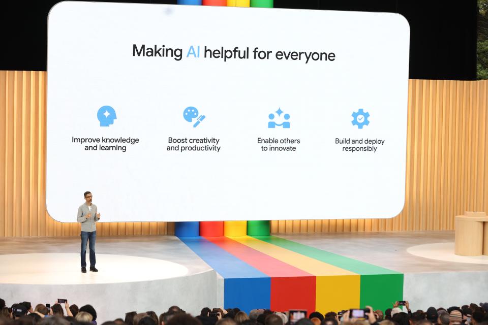 Sundar Pichai, in a grey jumper and jeans, stands on a stage with a rainbow-colored adornment in the center, as a screen displays "Making AI helpful for everyone"