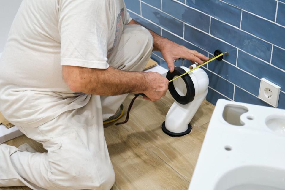 A person in white uses a tape measure while replace a toilet flange.