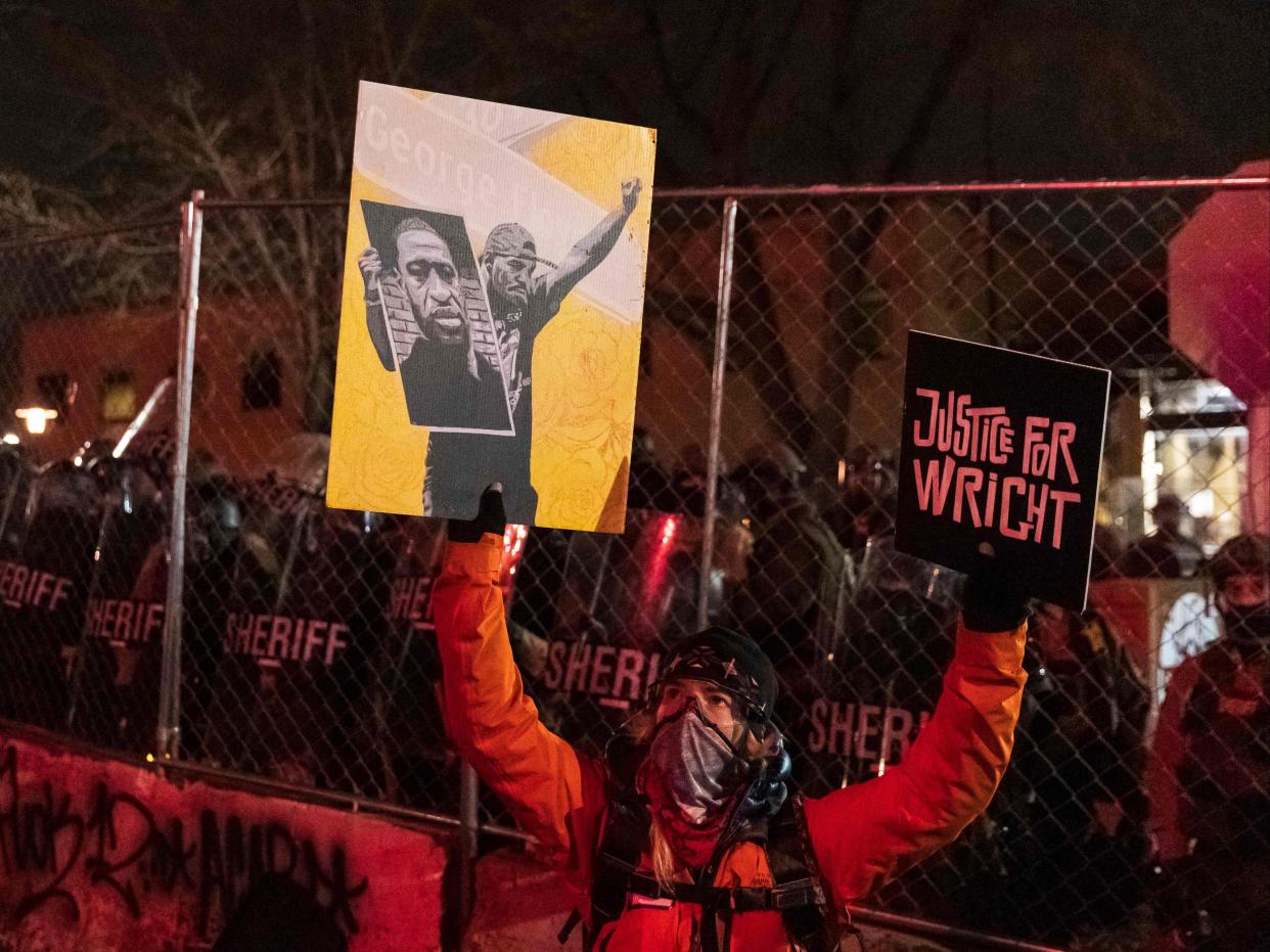 <p>A demonstrator holding a poster of George Floyd and sign reading “Justice for Wright” in front of a line of police officers outside the Brooklyn Center police station while protesting the death of Daunte Wright who was shot and killed by a police officer in Brooklyn Center, Minnesota on 14 April 2021</p> ((AFP via Getty Images))