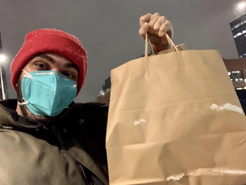 The writer (wearing a red beanie and blue mask) holding up a brown bag of Alinea takeout