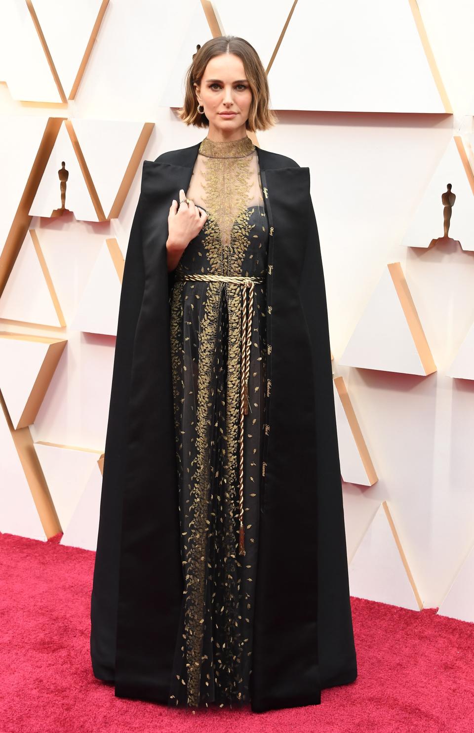 Natalie Portman in a black and gold dress and black cape on a red carpet