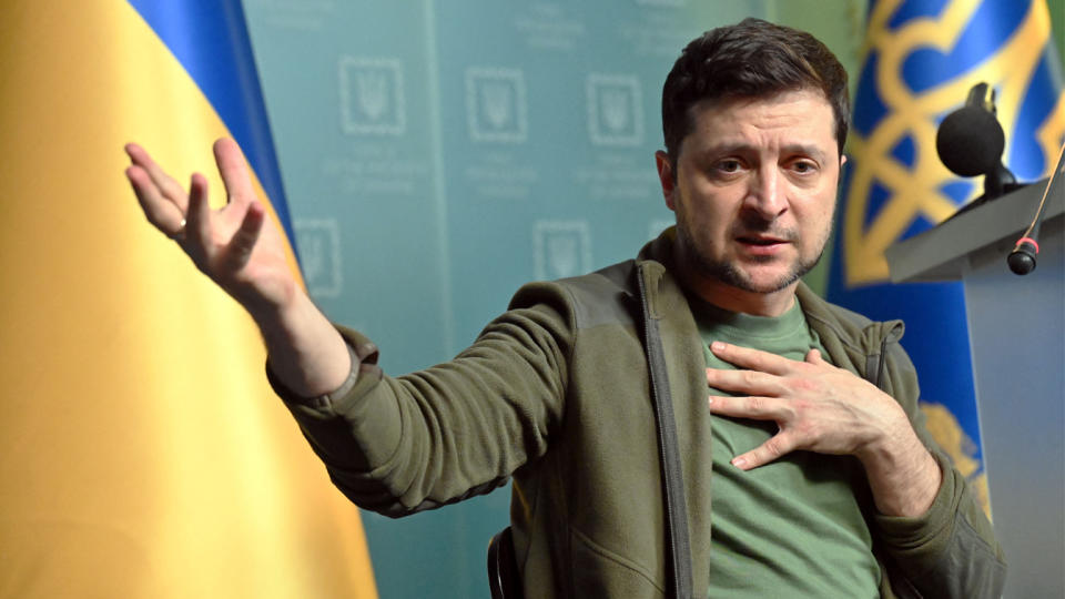 Ukrainian President Volodymyr Zelensky gestures, with his hand on his heart, as he addresses a microphone, with the Ukrainian blue and yellow flag in the background.