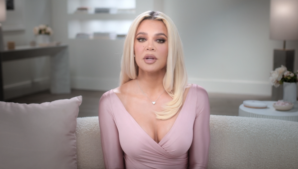 Khloé Kardashian sits on a couch in a stylish light pink dress, speaking directly to the camera in a scene from "The Kardashians" on Disney+