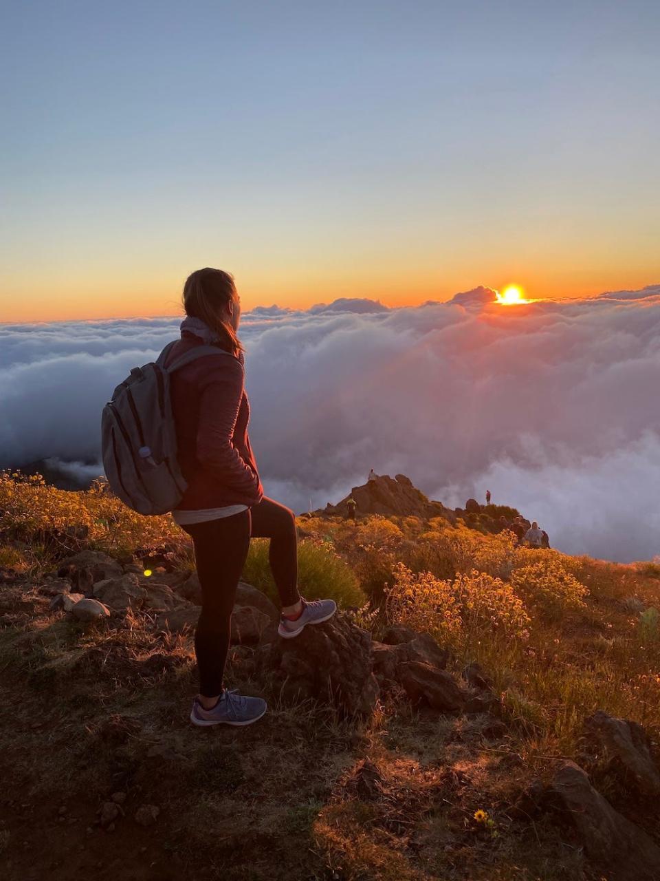 A woman facing away from the camera on top of a mountain. The sun rises through the clouds in front of her.