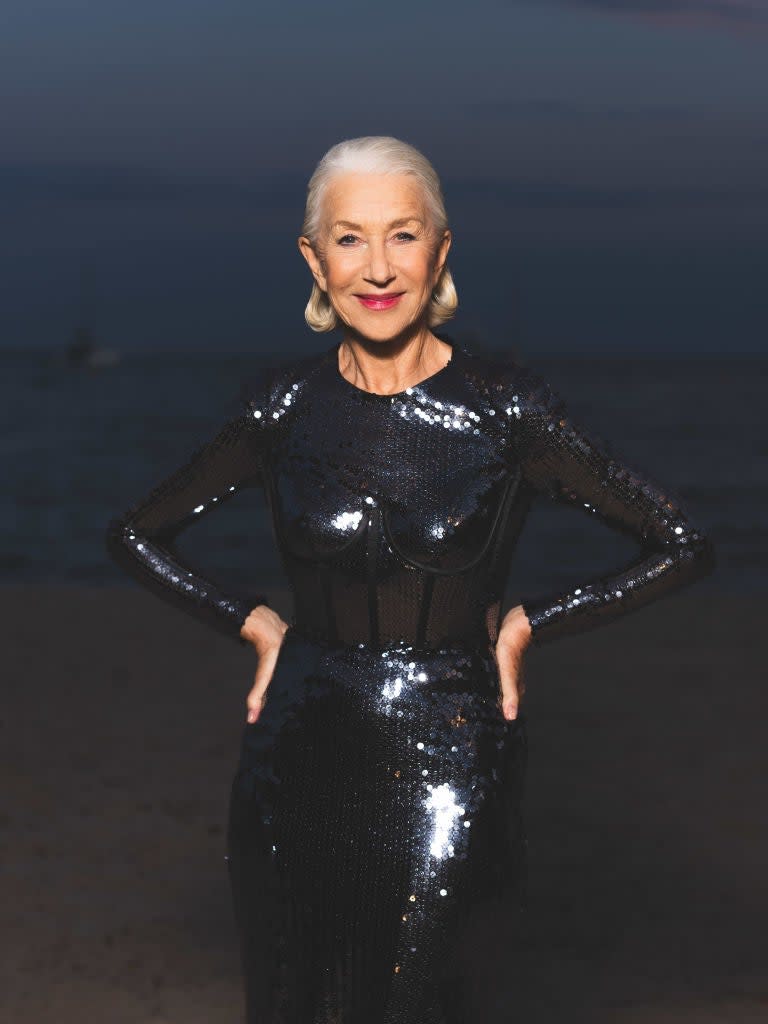 Helen Mirren on the beach, wearing a form-fitting, long-sleeved, sequined dress with her hands on her hips