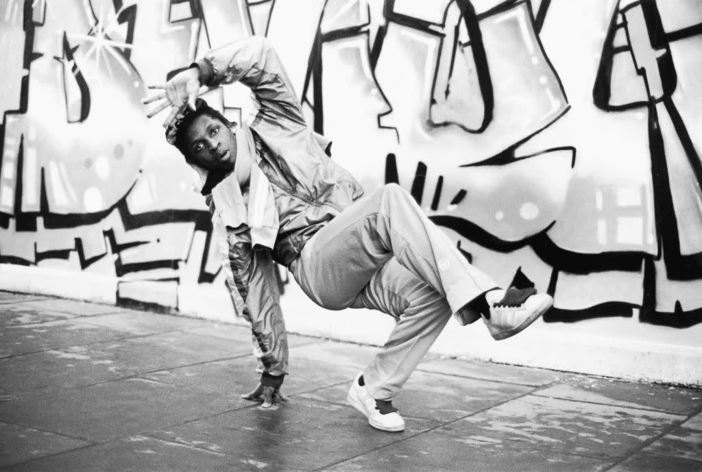 Check out these 60+ 80s trivia questions. Pictured: a black and white image of a man breakdancing. | A breakdancer in action against a graffiti backdrop,  Covent Garden, London, 1985. (Photo by Leon Morris/Hulton Archive/Getty Images)