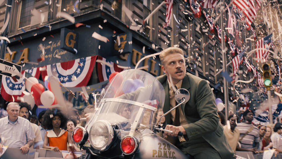 Still from the movie Indiana Jones and the Dial of Destiny. A man with short blond hair and wearing a suit is riding a New York police motorcycle, searching through a parade. There are American flags hanging everywhere and red, white and blue confetti and streamers about.