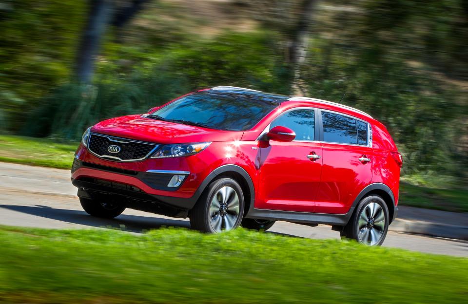 the front and left side of a red 2013 kia sportage driving on a road with grass in the foreground and trees in the background
