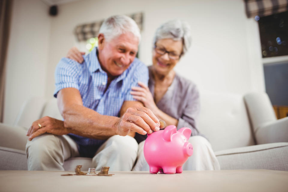 An older man sitting next to an older woman puts a coin into a piggy bank