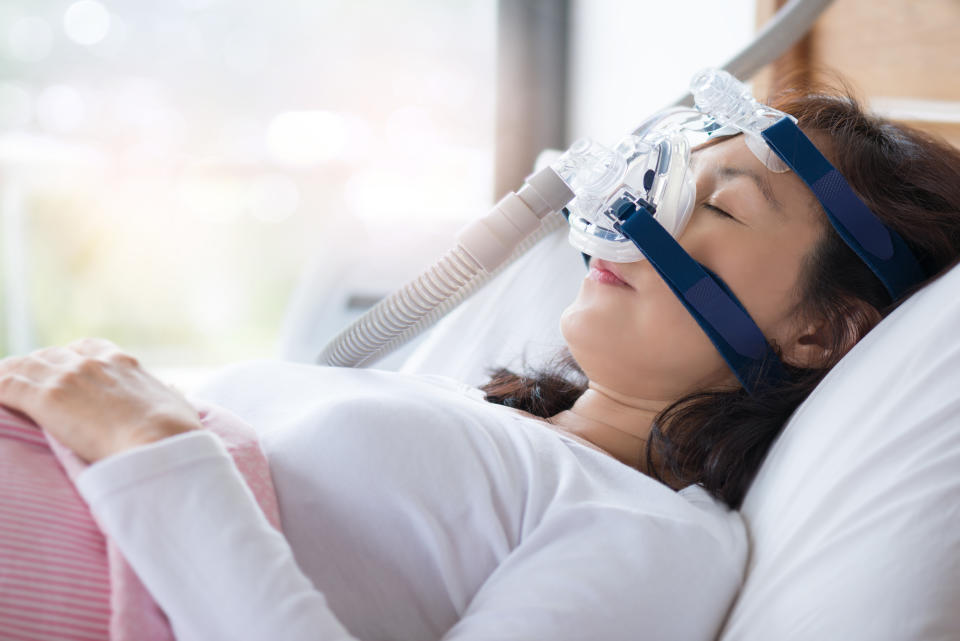 Senior woman using cpap machine to stop choking and snoring from obstructive sleep apnea with bokeh and morning light background.