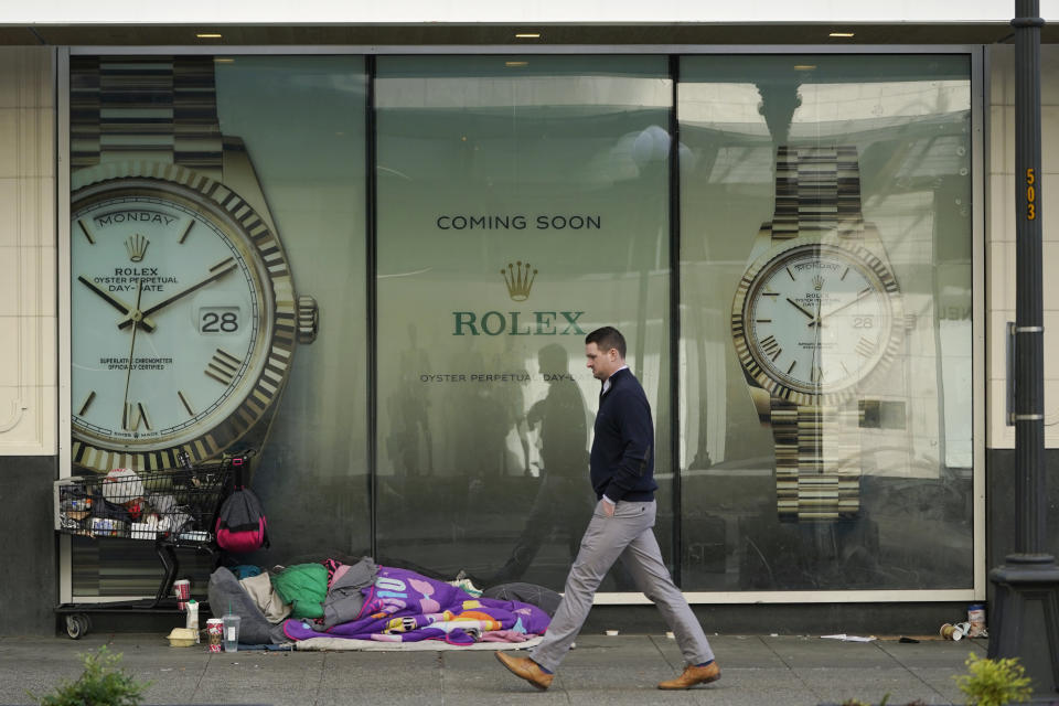 FILE - A person sleeps next to a shopping cart as a pedestrian walks past a store-window sign advertising the future opening of a Rolex watch store on Jan. 31, 2022, in downtown Seattle. In Feb. 2022, the mayor of Portland, Ore., banned camping on the sides of certain roadways, and officials are exploring other aggressive options to combat homelessness. In an increasing numbers of liberal cities like Portland, Seattle and New York, officials are cracking down on encampments after years of tolerating growing numbers of people living in tents. (AP Photo/Ted S. Warren, File)