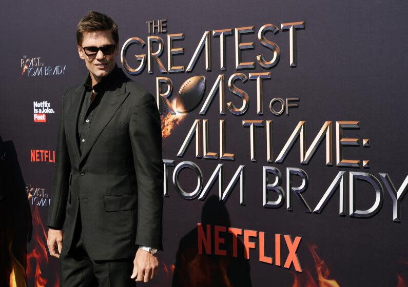 Tom Brady wears shades while standing in front of a sign reading "The Greatest Roast of All Time: Tom Brady"