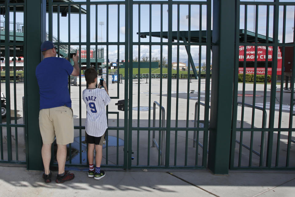 FILE - Cubs fans take photos through the locked gates at Sloan Park, the spring training site of the Chicago Cubs, in Mesa, Ariz., after Major League Baseball suspended the rest of its spring training game schedule because of the coronavirus outbreak, in this Friday, March 13, 2020, file photo. The Cactus League and Arizona community leaders have asked Major League Baseball to delay the start of spring training due to coronavirus concerns just over three weeks before pitchers and catchers are supposed to report. The Cactus League made the request in a letter to Baseball Commissioner Rob Manfred obtained by The Associated Press on Monday, Jan. 25, 2021. (AP Photo/Sue Ogrocki, File)