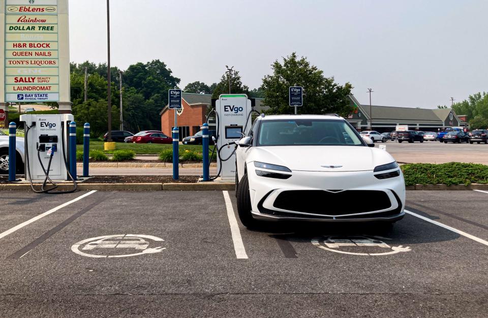 A white Genesis GV60 SUV charges at an EVgo charging station, with an overcast sky seen in the background.