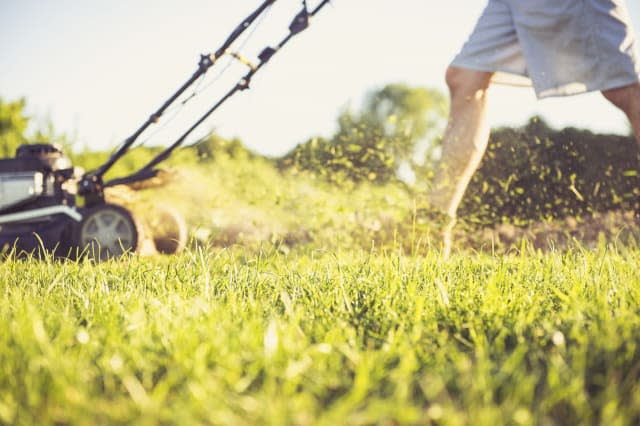 Low section of man mowing grassy field in yard against sky during sunset