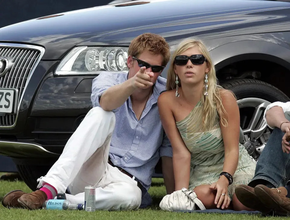 prince harry pointing at the camera as he sits next to Chelsy Davy.