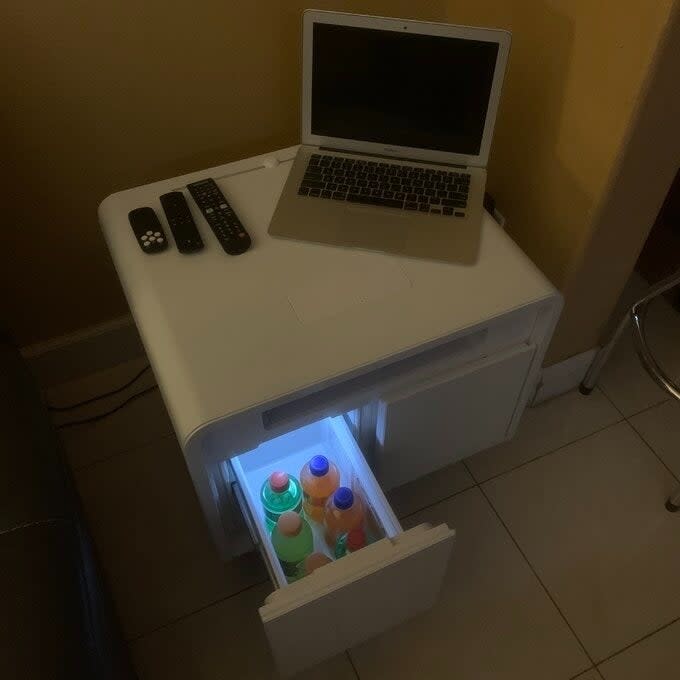 a reviewer photo of the white nightstand with an open drawer showcasing drinks, beside a laptop and remotes on top