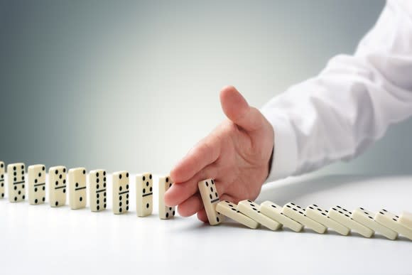 Businessman's hand stops a row of dominoes from falling, connoting a successful defensive strategy.