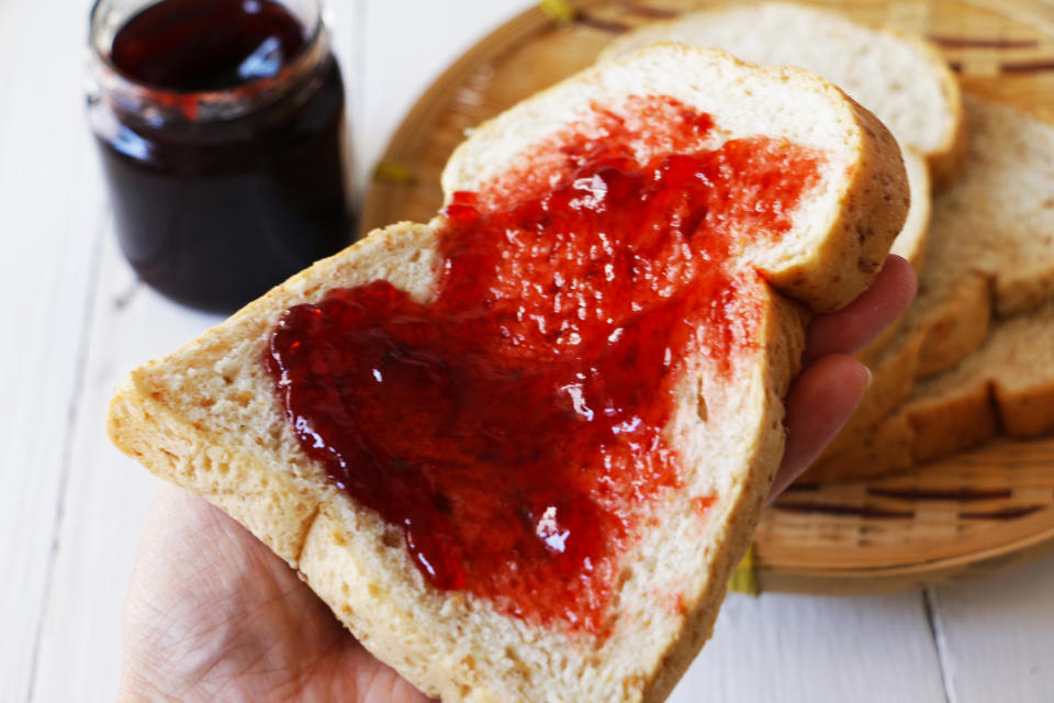 A hand holds a slice of bread spread with strawberry jam, a jar of jam in the background on a wooden tray