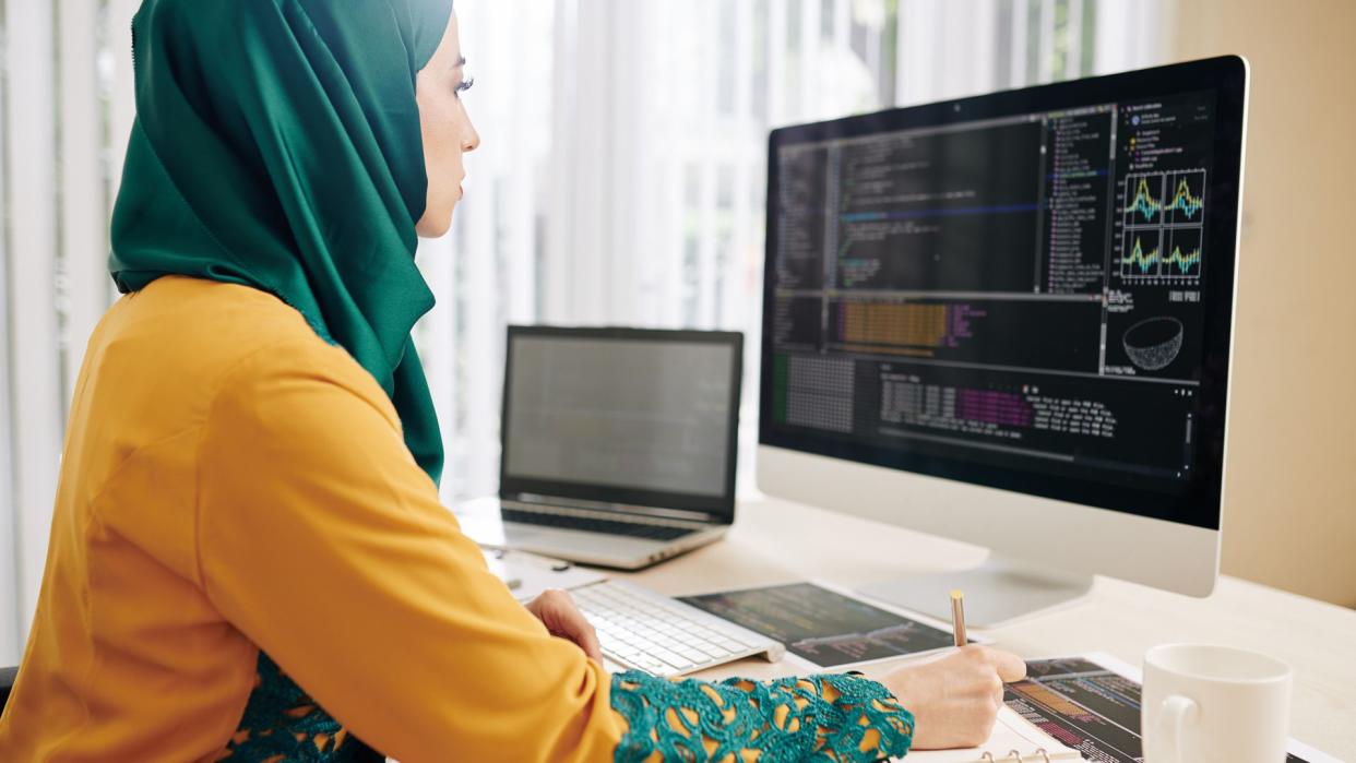 Young woman in hijab checking code implementation and taking notes in planner.