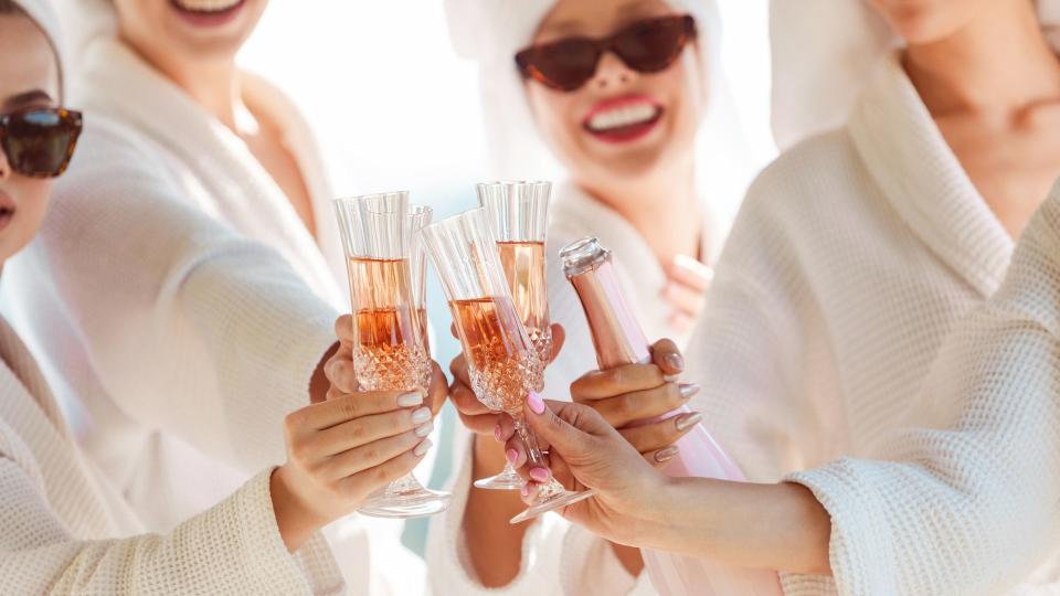 female friends in white bathrobes before a bachelorette party toasting with champagne