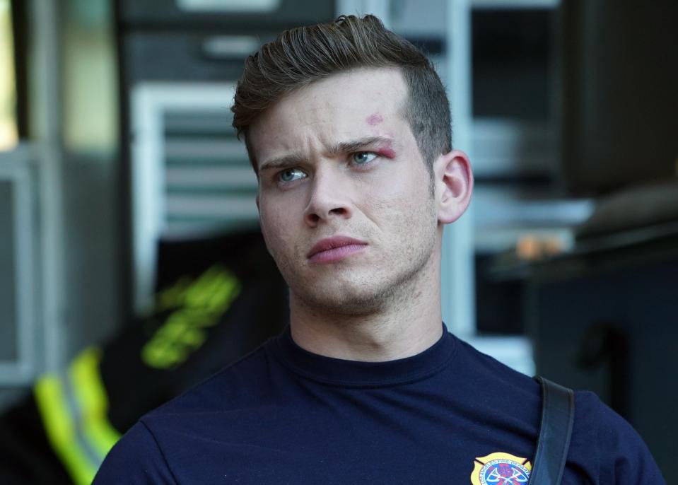 TV show character in firefighter attire sitting pensively beside a firetruck