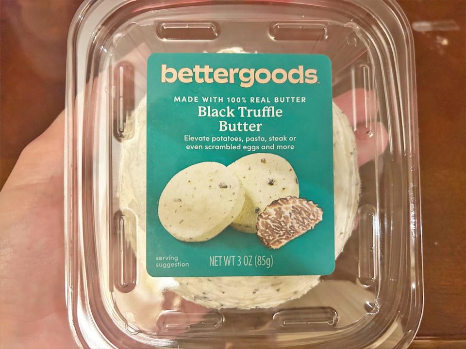 A hand holds a clear plastic container of butter with a blue label with "Bettergoods black truffle butter" text on it