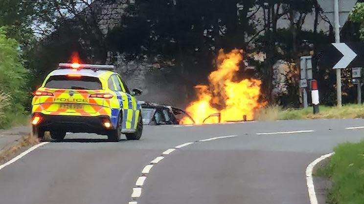 A police car parked on a road near Ingham with a vehicle in flames in the background. 