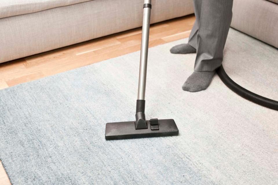 A person in grey pants and socks pushes a vacuum across a grey carpet.
