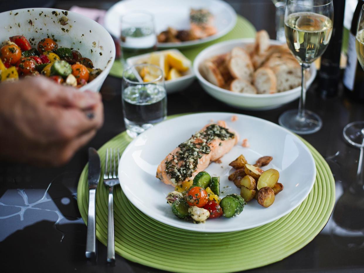 A plate of salmon and veggies, with a glass of wine, bread, and salad in the background — a Mediterranean diet meal