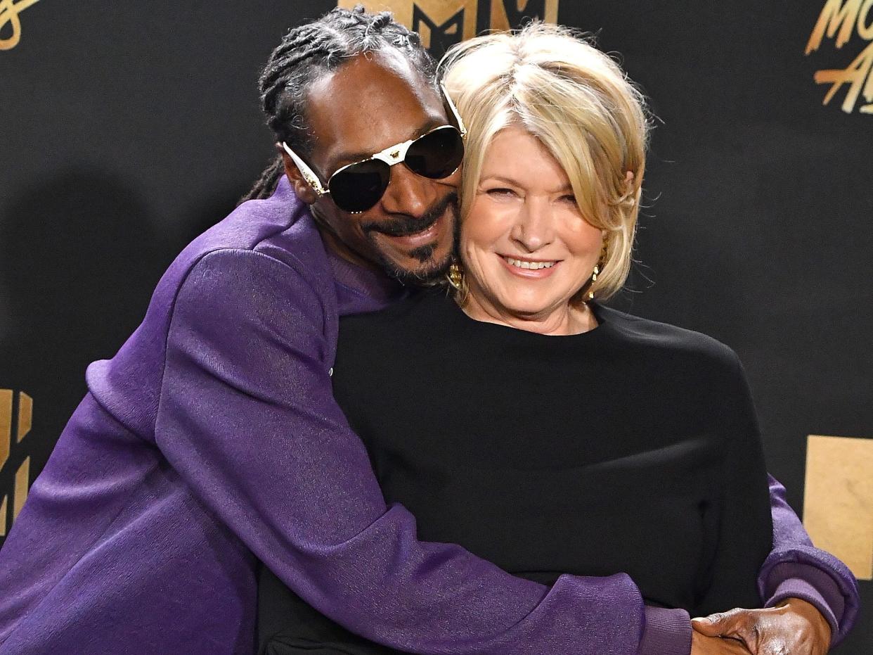 Snoop Dogg, who is wearing a purple jacket and a pair of blacked-out sunglasses, hugging Martha Stewart, who is wearing a black top.