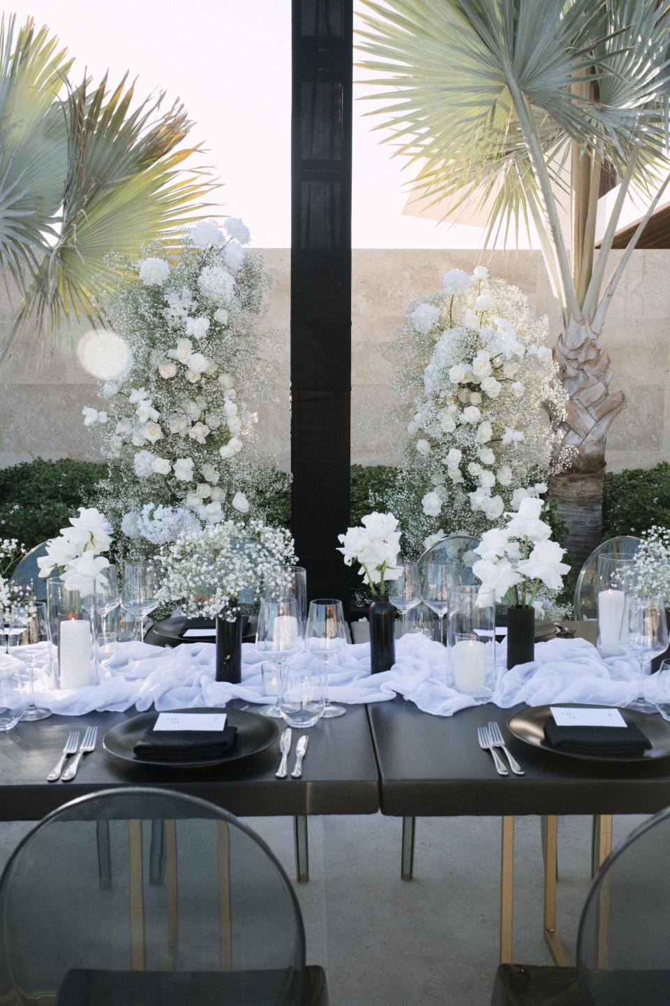A black table with a white runner, black vases, and white flowers. A floral arch sits behind it. 