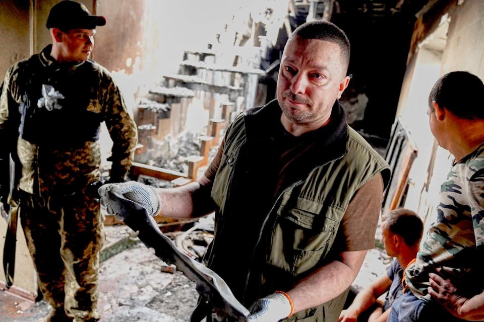 A man with close-cropped hair and a grim expression holds a large fragment with gloved hands in a debris-covered room