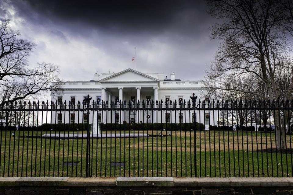 A large white mansion with dark clouds over it, behind a fence.