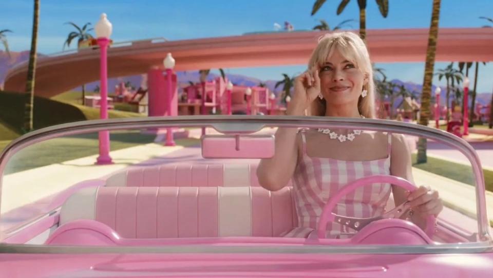 Margot Robbie's Barbie smiles in her pink dress driving her pink cadillac in pink Barbieland