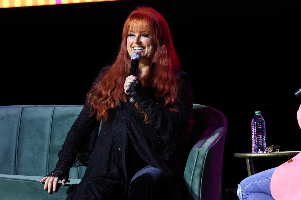 Wynonna Judd sits in chair and holds microphone