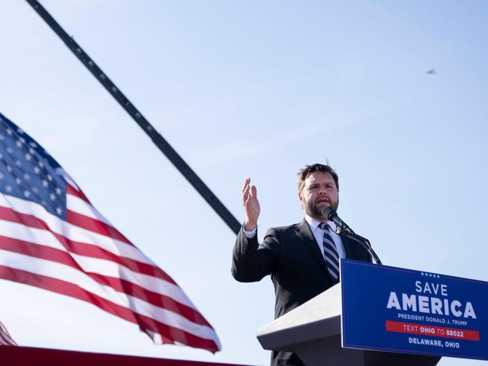 A man standing at a podium outside with the American flag hanging in the background.