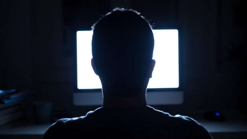 An unseen man sits in front of a computer screen at night.