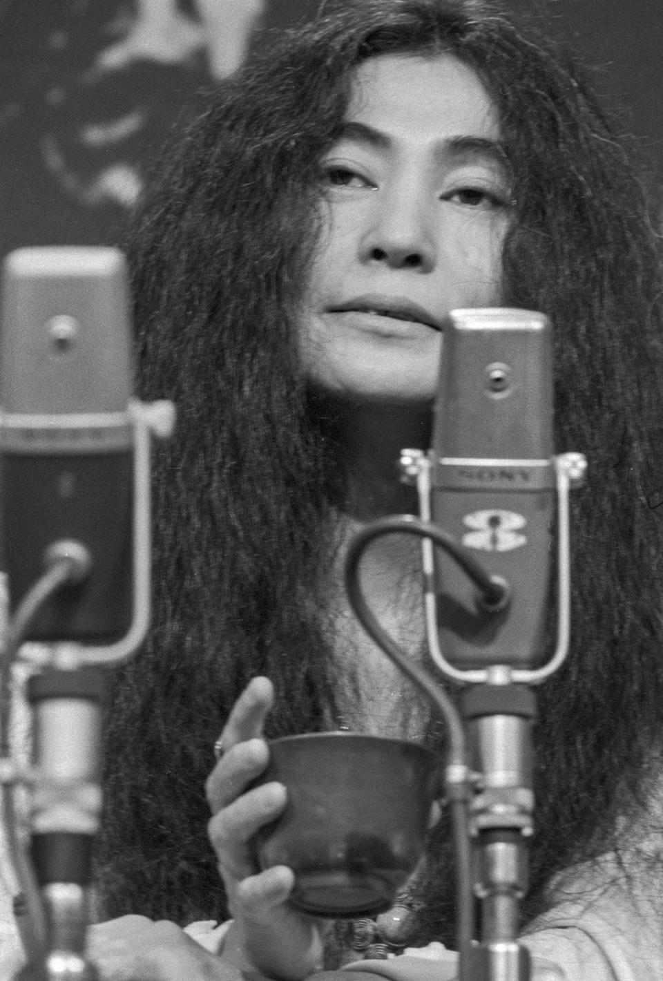 Photo of Yoko Ono in front of a microphone holding a cup of tea