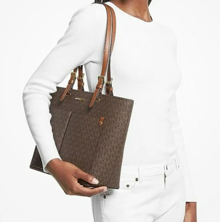 Person holding a brown designer tote bag with a white long-sleeve top and white pants